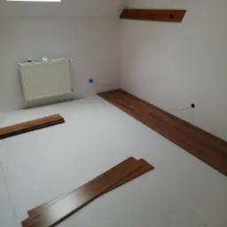 Poddasze z oknem dachowym, przygotowane do montażu paneli podłogowych, widoczna warstwa izolacyjna i ułożone deski paneli w kolorze drewna.