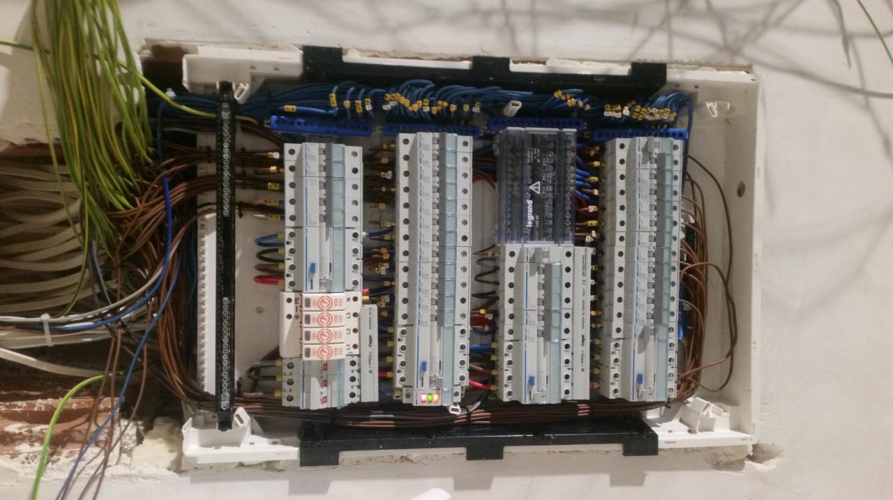 Rozdzielnica elektryczna w trakcie montażu w osadzonej w ścianie obudowie, widoczne liczne kable, bezpieczniki, moduły i oznaczenia, z jednym świecącym zielonym LED, w tle surowa ściana.