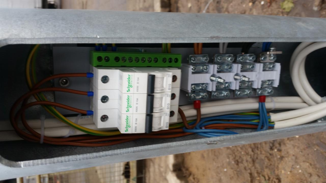 Wnętrze metalowej skrzynki rozdzielczej z zainstalowanymi bezpiecznikami Schneider Electric, zieloną listwą zaciskową i białymi złączkami, widoczne przewody w różnych kolorach zabezpieczone...