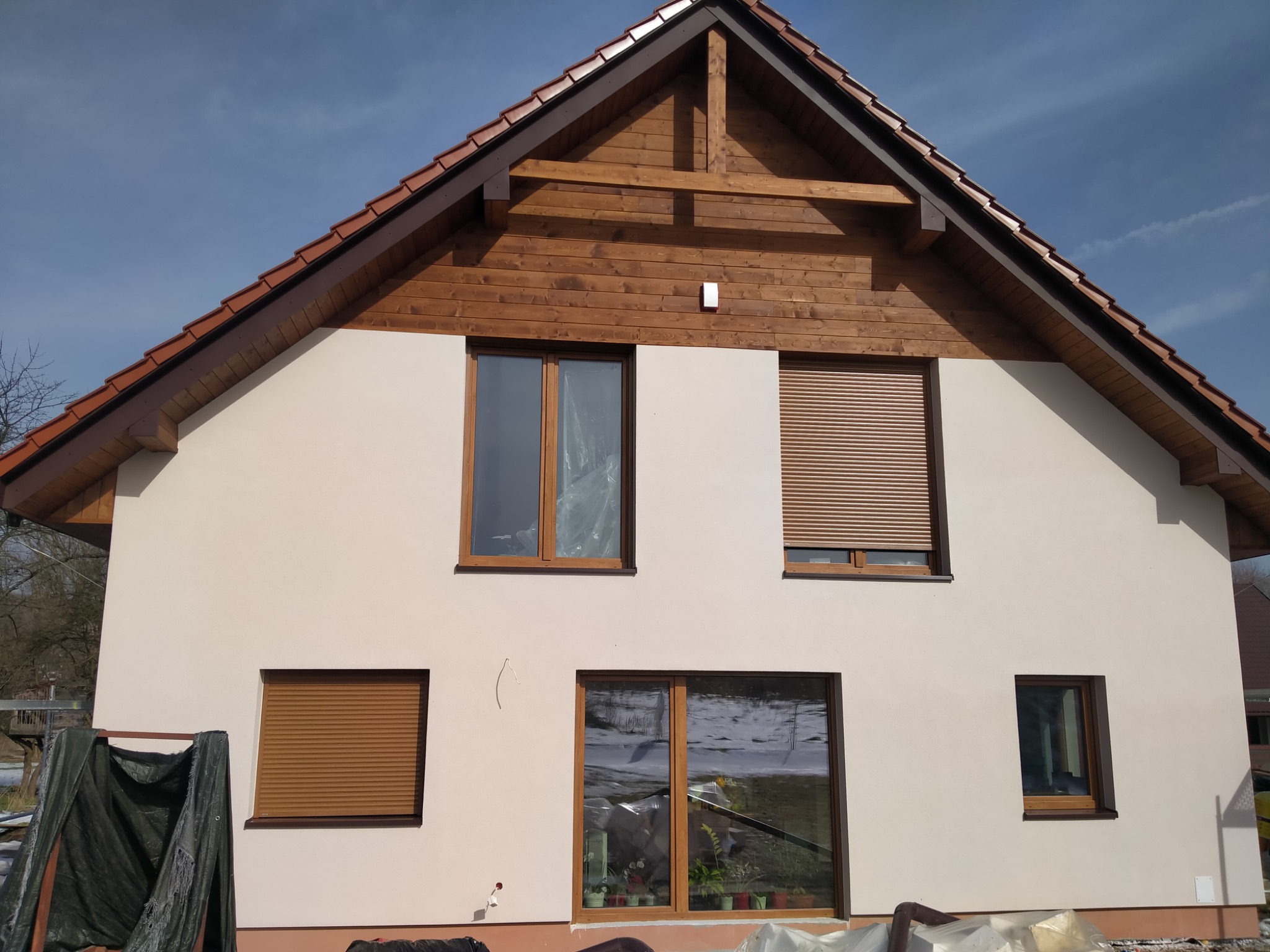 Dwupiętrowy dom z jasną elewacją, drewnianą konstrukcją szczytową i brązowymi roletami na niektórych oknach, częściowo zasłonięty folią ochronną i plandeką.