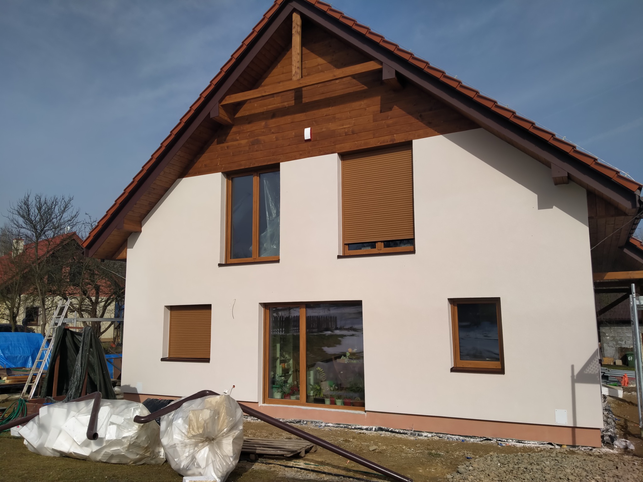 Świeżo otynkowana elewacja domu jednorodzinnego z brązowymi oknami i roletami, widoczne worki z materiałem izolacyjnym i rusztowanie w tle.