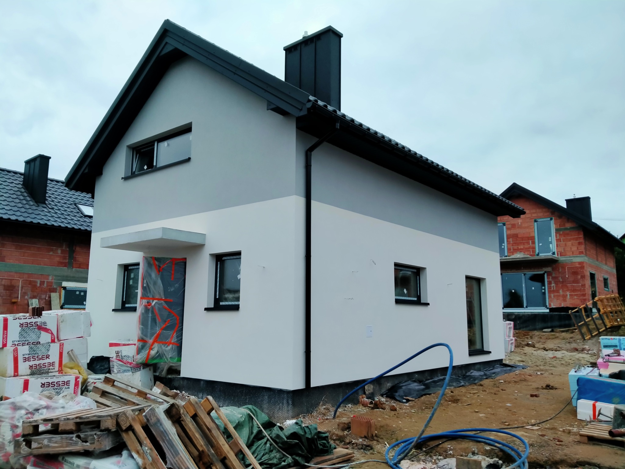 Nowoczesny dom jednorodzinny z szarą górną i białą dolną częścią elewacji, czarnym dachem i orynnowaniem, widoczne materiały budowlane i narzędzia sugerują prace wykończeniowe elewacji.