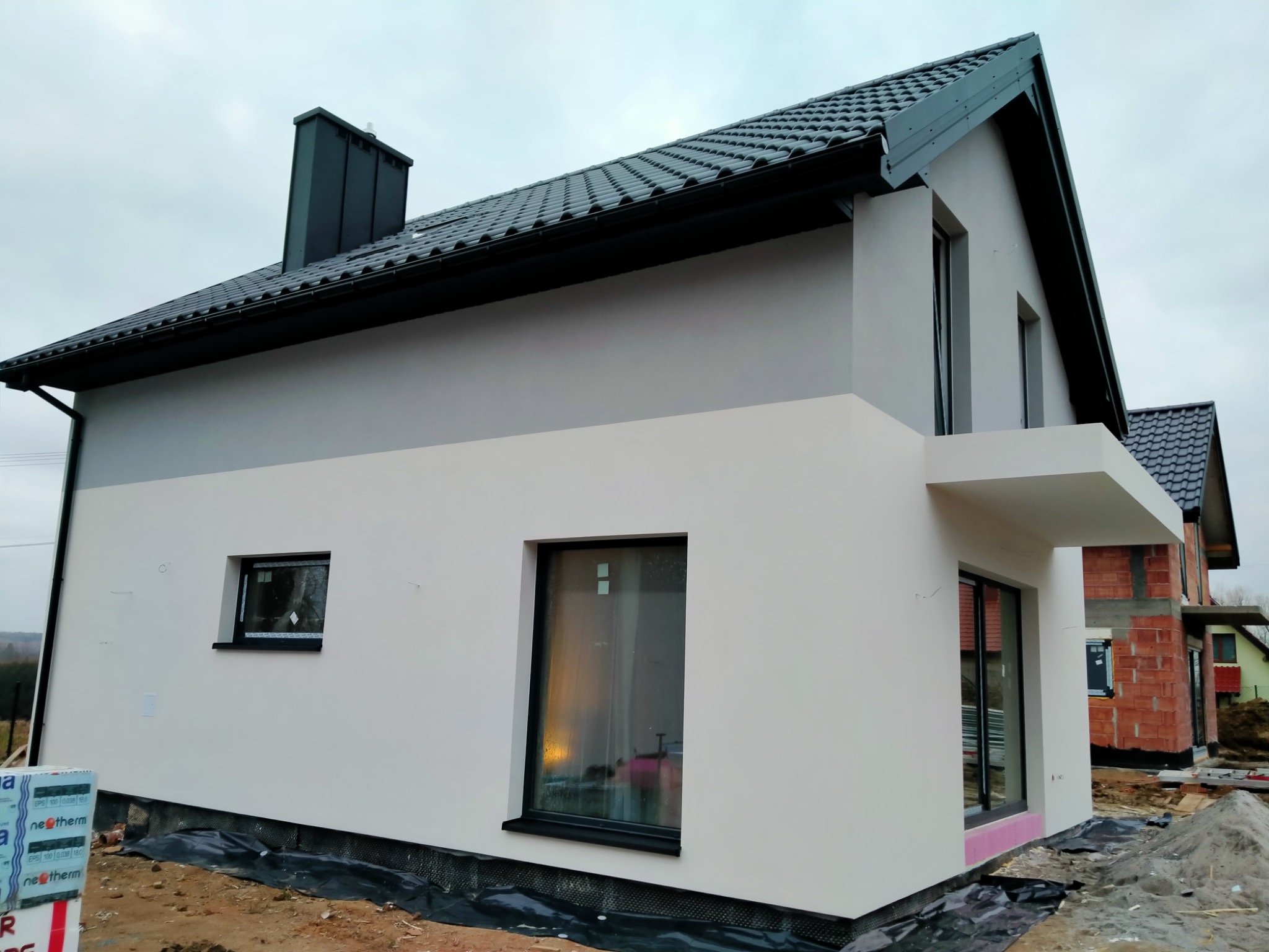 Dwukondygnacyjny dom jednorodzinny z szaro-białą elewacją, czarną dachówką i oknami, w trakcie prac wykończeniowych, widoczne materiały izolacyjne i fragment budynku w surowej cegle
