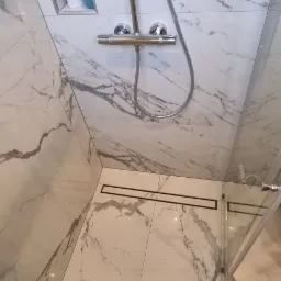 Nowoczesna kabina prysznicowa z marmurowymi płytkami, chromowaną armaturą i odpływem liniowym, uzupełniona o turkusowy dywanik.