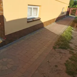 Elewacja domu z żółtym tynkiem, brązowym cokołem i nową kostką brukową na ścieżce, widok z boku w słoneczny dzień.