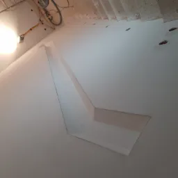 Świeżo pomalowany sufit z nietypowym kształtem, z widocznymi śladami po otworach na oświetlenie i kablem elektrycznym w rogu, oświetlony lampą halogenową na statywie.