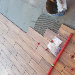 Układanie płytek imitujących drewno na podłodze, widoczna warstwa kleju, poziomica oraz system poziomowania płytek z czerwonymi klipsami i białymi krzyżykami.