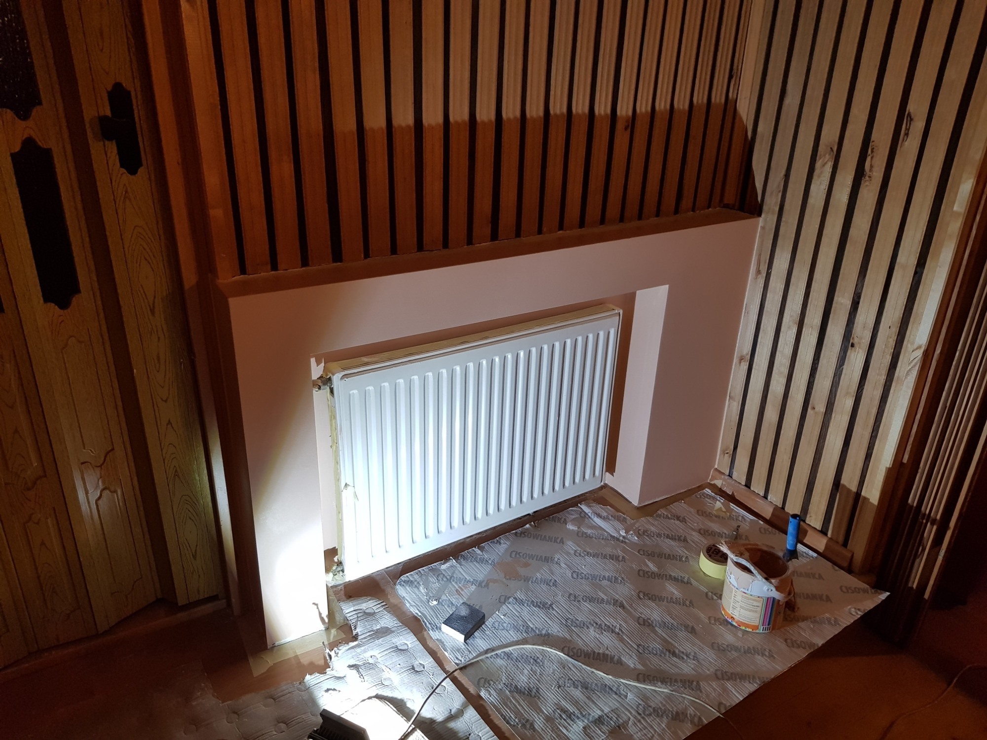 Przygotowanie do malowania ściany z grzejnikiem: zabezpieczona folią podłoga, puszka z farbą, wałek i szpachelka, ściana w kolorze pudrowego różu, drewniana boazeria pionowa