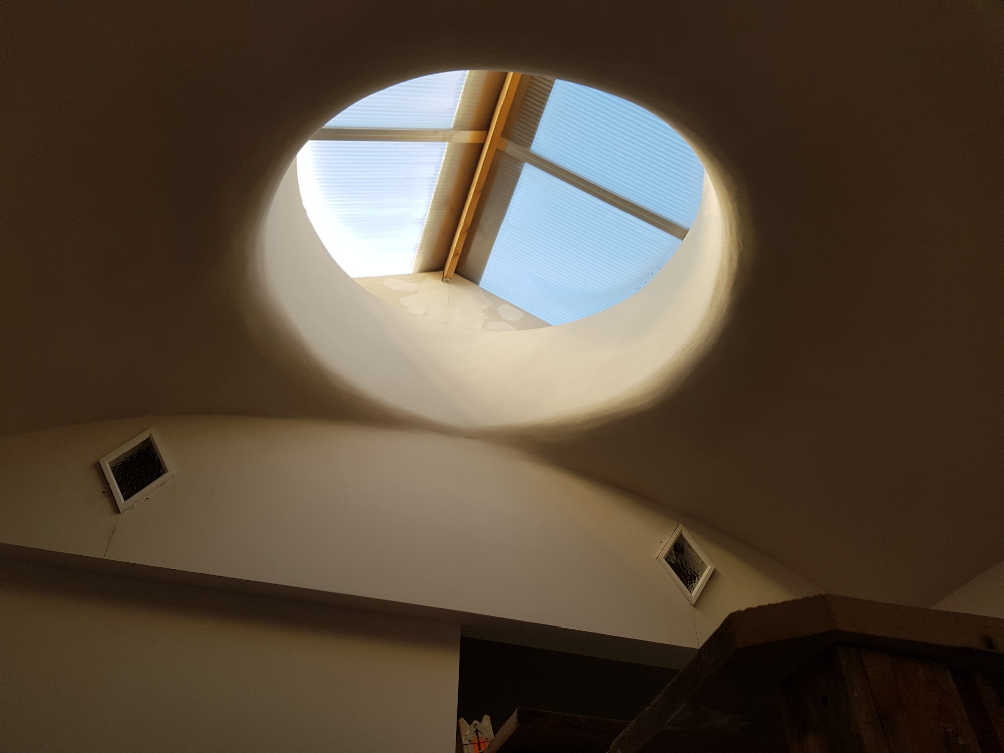 Okrągłe okno dachowe z widoczną konstrukcją drewnianą i niebieskim niebem, umieszczone w białym suficie z delikatnym oświetleniem i małymi kratkami wentylacyjnymi po bokach.