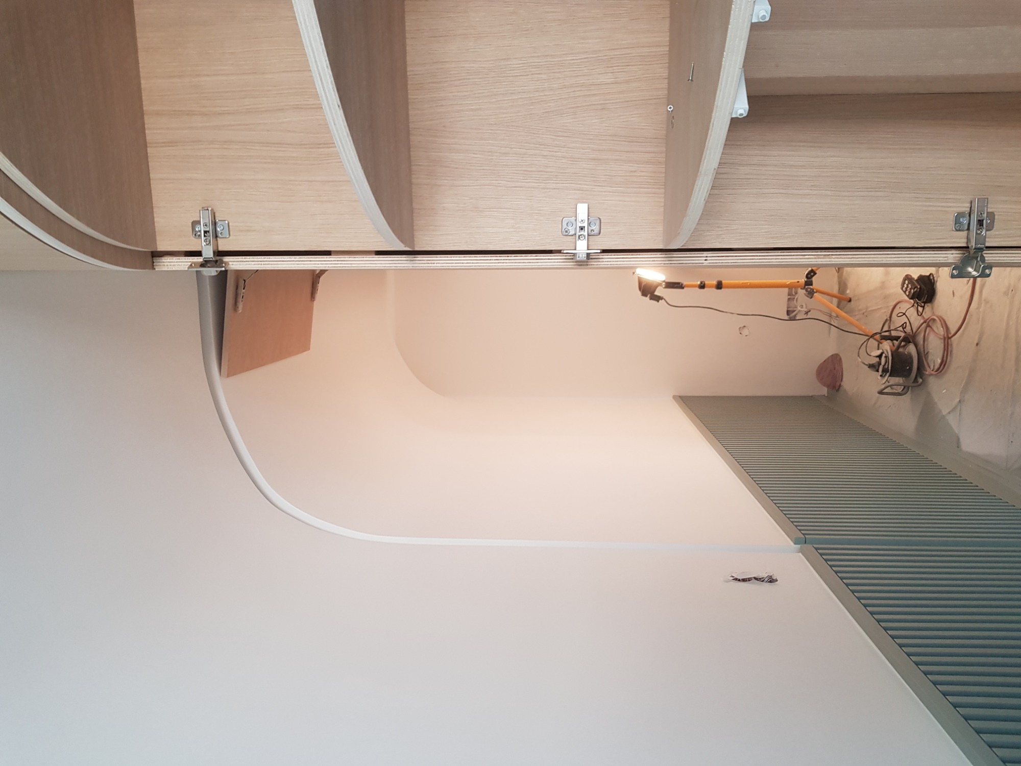 Montaż drewnianych półek z zaokrąglonymi krawędziami i metalowymi zawiasami, widoczna lampa robocza na trójnogu i fragment ściany z żaluzjami w trakcie prac wykończeniowych.