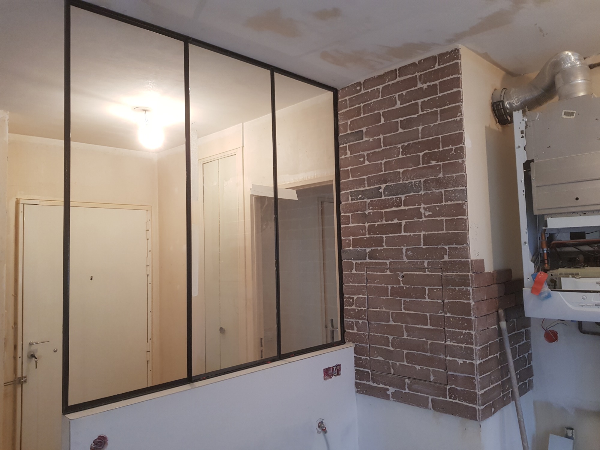 Wnętrze w trakcie prac wykończeniowych: szklana ścianka działowa z czarnymi profilami oddziela pomieszczenia, obok ściana wyłożona cegłą klinkierową w odcieniach brązu, widoczny piec gazowy...