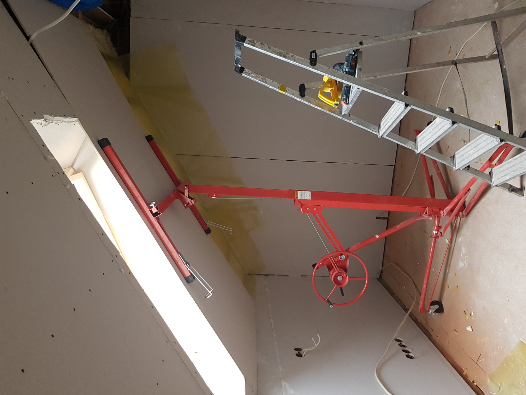 Montaż płyty kartonowo-gipsowej na poddaszu z użyciem podnośnika i drabiny, widoczne otwory na wkręty i przewody elektryczne