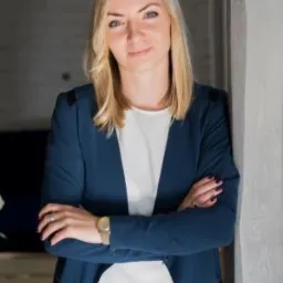 Portret uśmiechniętej blondynki w granatowej marynarce, opierającej się o ścianę, z założonymi rękami i zegarkiem na nadgarstku.