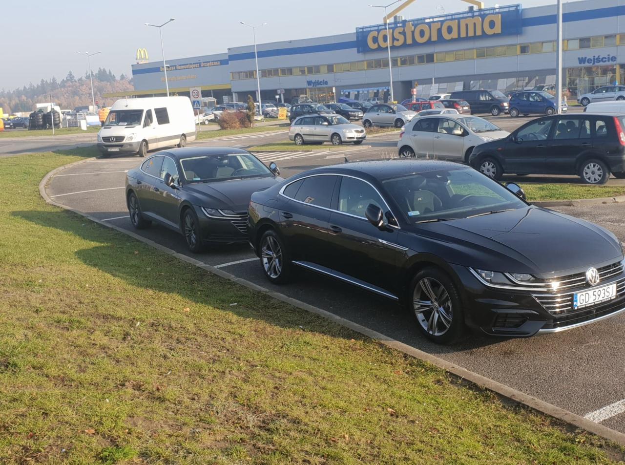 Dwa czarne samochody Volkswagen Arteon zaparkowane na parkingu przed sklepem Castorama w słoneczny dzień.