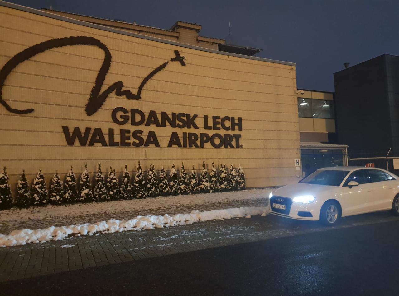 Białe Audi A3 z włączonymi światłami zaparkowane przed budynkiem lotniska Gdańsk im. Lecha Wałęsy, zimowy krajobraz z małymi choinkami i śniegiem.