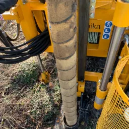 Żółta wiertnica geologiczna w trakcie wiercenia otworu w ziemi, widoczny fragment kolumny wiertniczej z wyraźnymi śladami ziemi i błota.