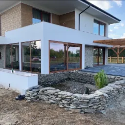 Nowoczesny dom z ogrodem w trakcie aranżacji: widoczny staw z kamienną obudową, taras wyłożony kostką brukową i drewniana pergola, ziemia wokół w trakcie prac.