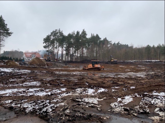 Teren przygotowywany pod budowę: pomarańczowy buldożer i dwie koparki na błotnistym placu z resztkami śniegu, w tle domy i las.
