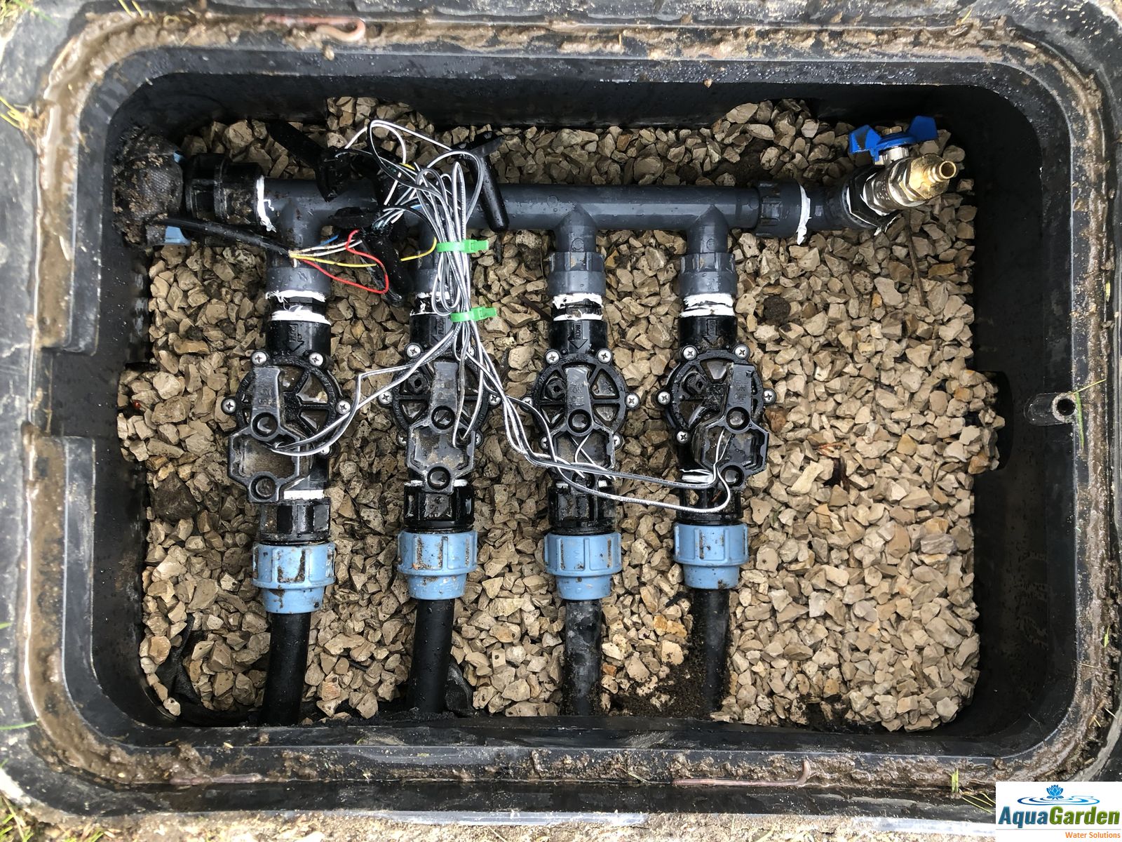 Skrzynka z czterema zaworami nawadniającymi, przewodami elektrycznymi i kamiennym wypełnieniem, widoczne z góry, z logo firmy AquaGarden w prawym dolnym rogu.