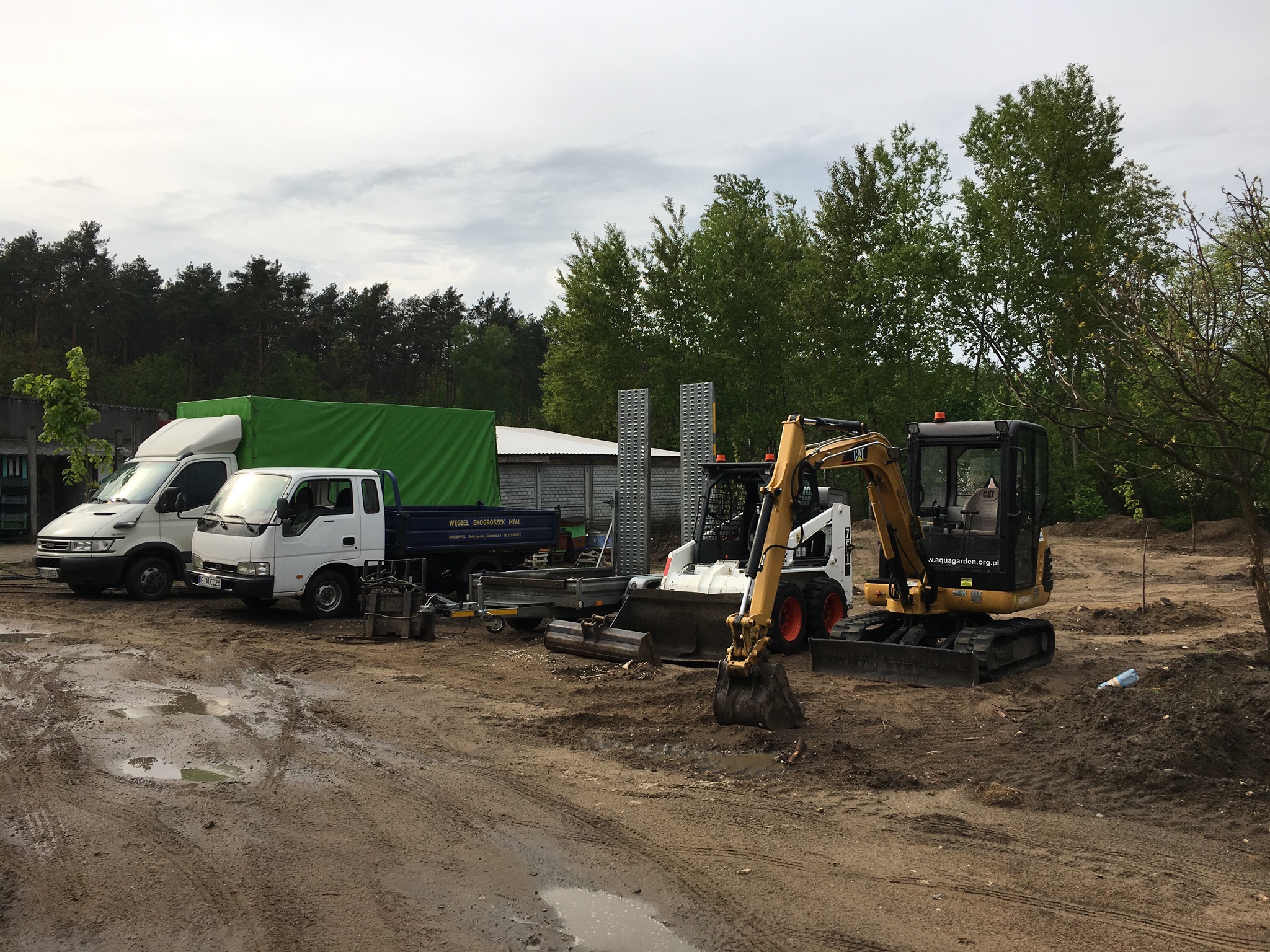 Park maszyn budowlanych: biały i żółty mini-ładowarki oraz wywrotka z przyczepą na błotnistym terenie, w tle zieleń drzew.