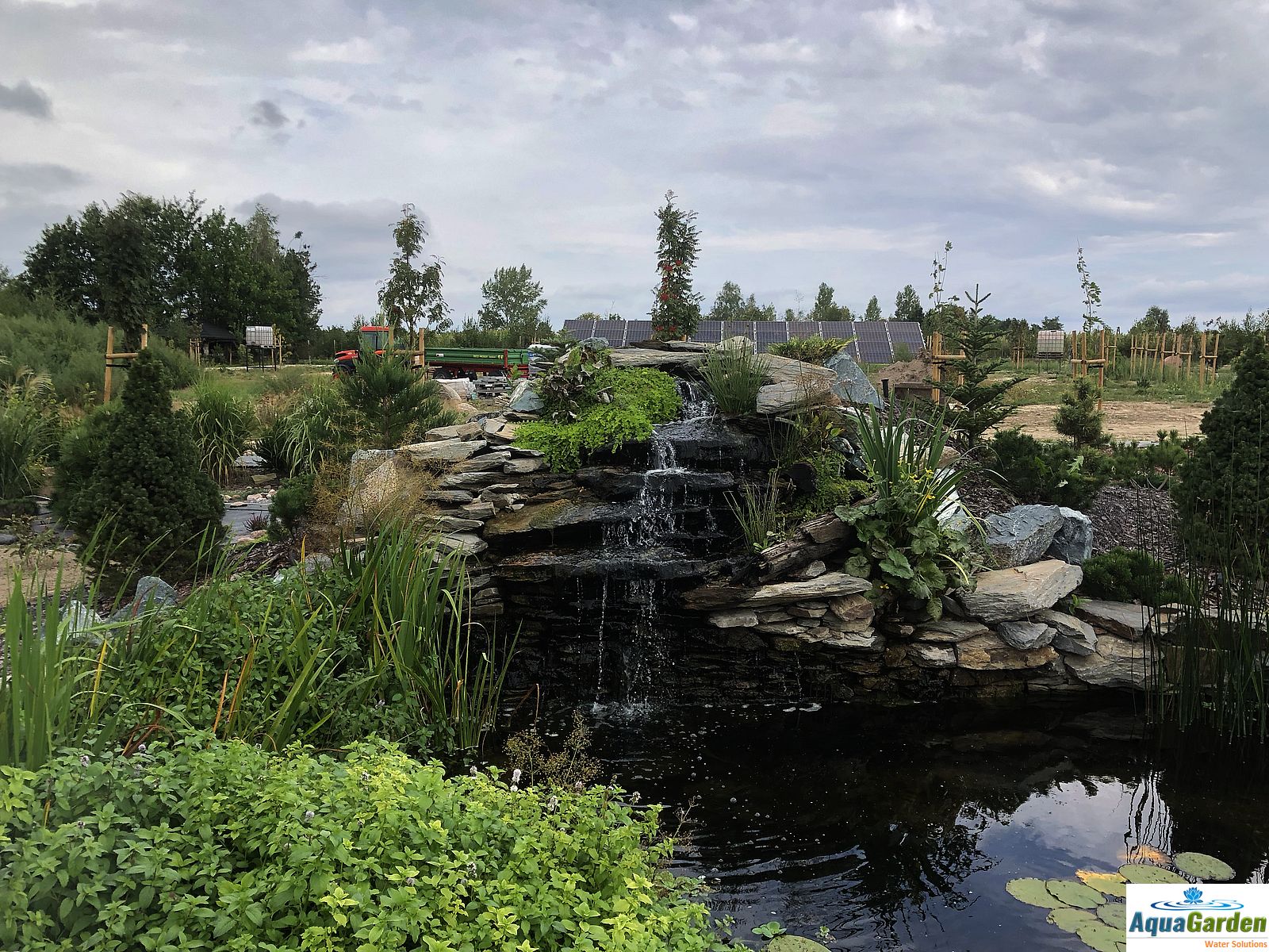 Kaskada wodna z kamieni w ogrodzie, otoczona zielenią, w tle widoczny traktor i panele słoneczne.