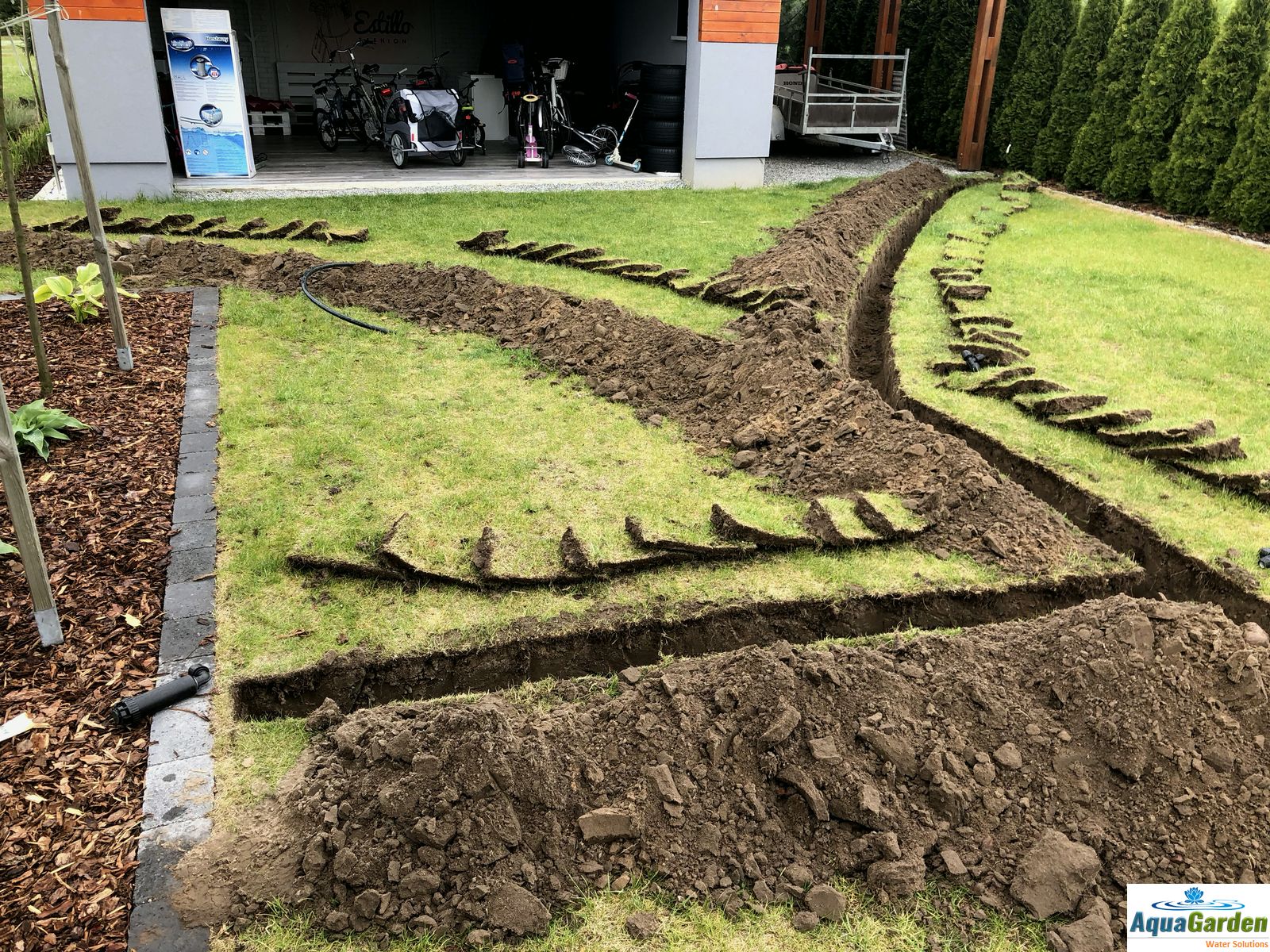 Wykopane rowy w trawniku pod drenaż, z odłożonymi darniami, widoczne rury i logo AquaGarden w rogu.