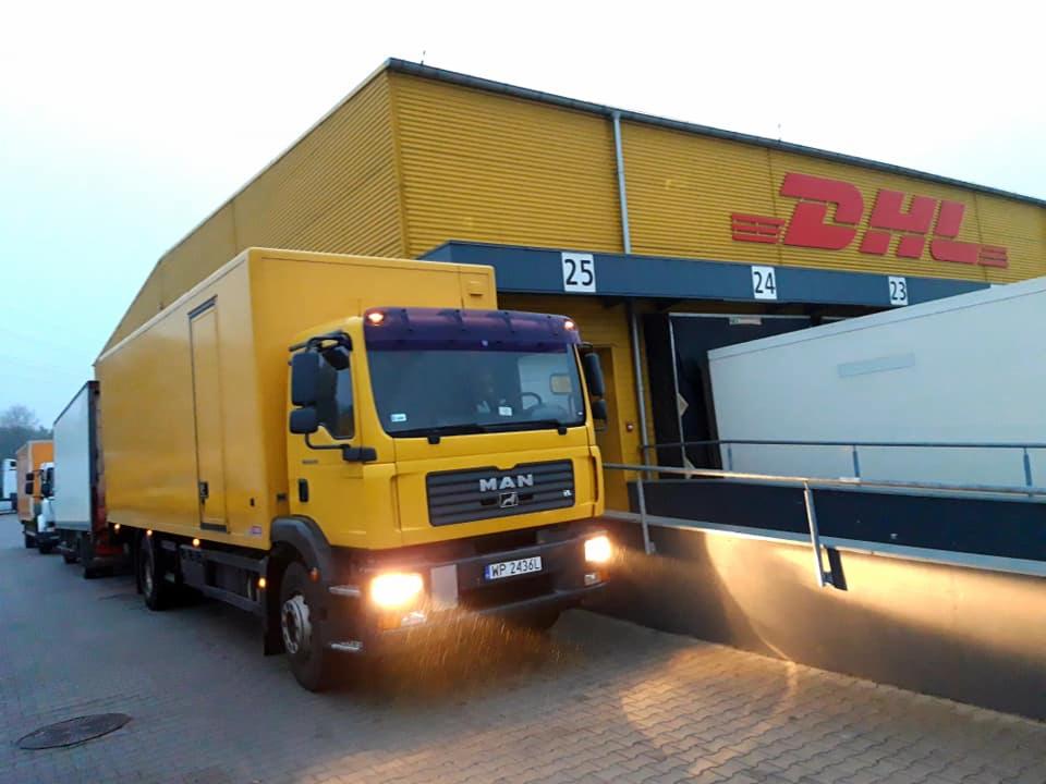 Żółta ciężarówka MAN z naczepą zaparkowana przy rampie załadunkowej DHL o numerze 25, z włączonymi światłami mijania, widoczna tablica rejestracyjna WP 2436L.