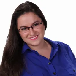 Uśmiechnięta kobieta w okularach i niebieskiej koszuli na białym tle, z długimi brązowymi włosami, patrzy w obiektyw.