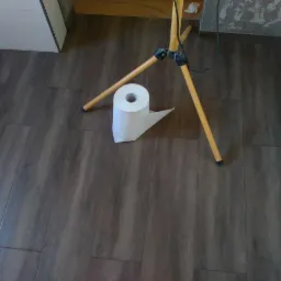 Ułożone panele podłogowe w odcieniach brązu, oświetlane lampą budowlaną na trójnogu. Na podłodze leży rolka papieru toaletowego.