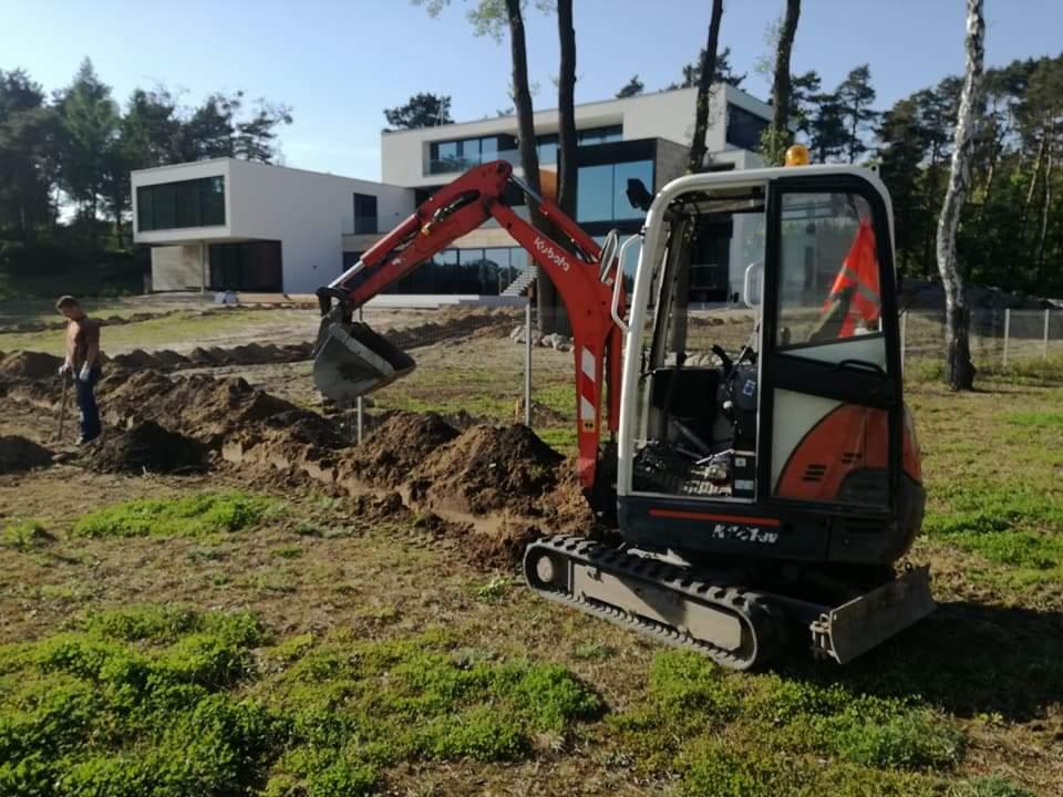 Mini koparka Kubota w trakcie wykonywania wykopu liniowego w gruncie, z widocznym operatorem z łopatą w tle i nowoczesnym domem jednorodzinnym w tle, w słoneczny dzień.