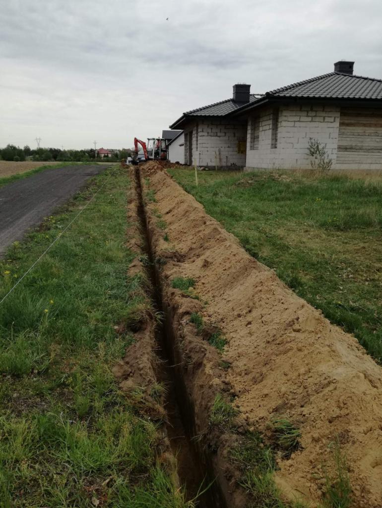 Długi, wąski wykop w ziemi, prawdopodobnie pod instalacje, z minikoparką widoczną w oddali na tle budynków w trakcie budowy.