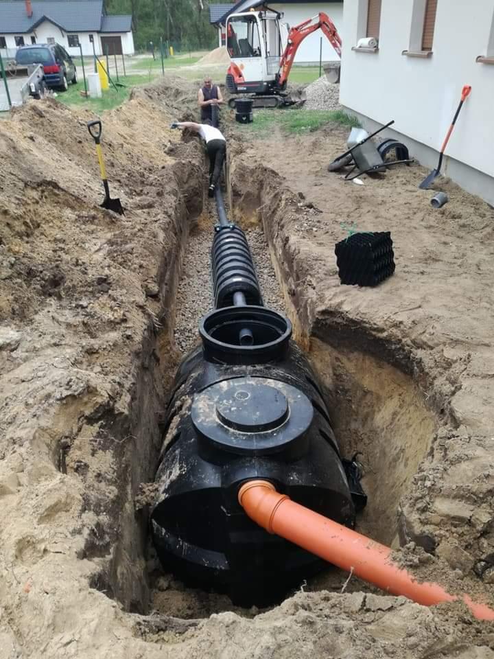 Czarny, plastikowy zbiornik na szambo zakopany w ziemi, widoczny pomarańczowy rurociąg, wykop otoczony ziemią, w tle minikoparka i pracownicy.