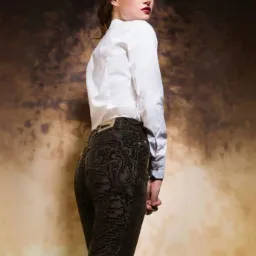 Kobieta odwrócona przez ramię, prezentująca ciemne spodnie z wytłaczanym wzorem paisley i białą koszulę, z logo marki w prawym górnym rogu na rozmytym tle.
