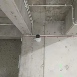 Kamera monitoringu zamontowana na betonowej ścianie z widocznymi przewodami elektrycznymi w białych i czerwonych rurkach. Widoczne zielone oznaczenia laserowe na ścianie.