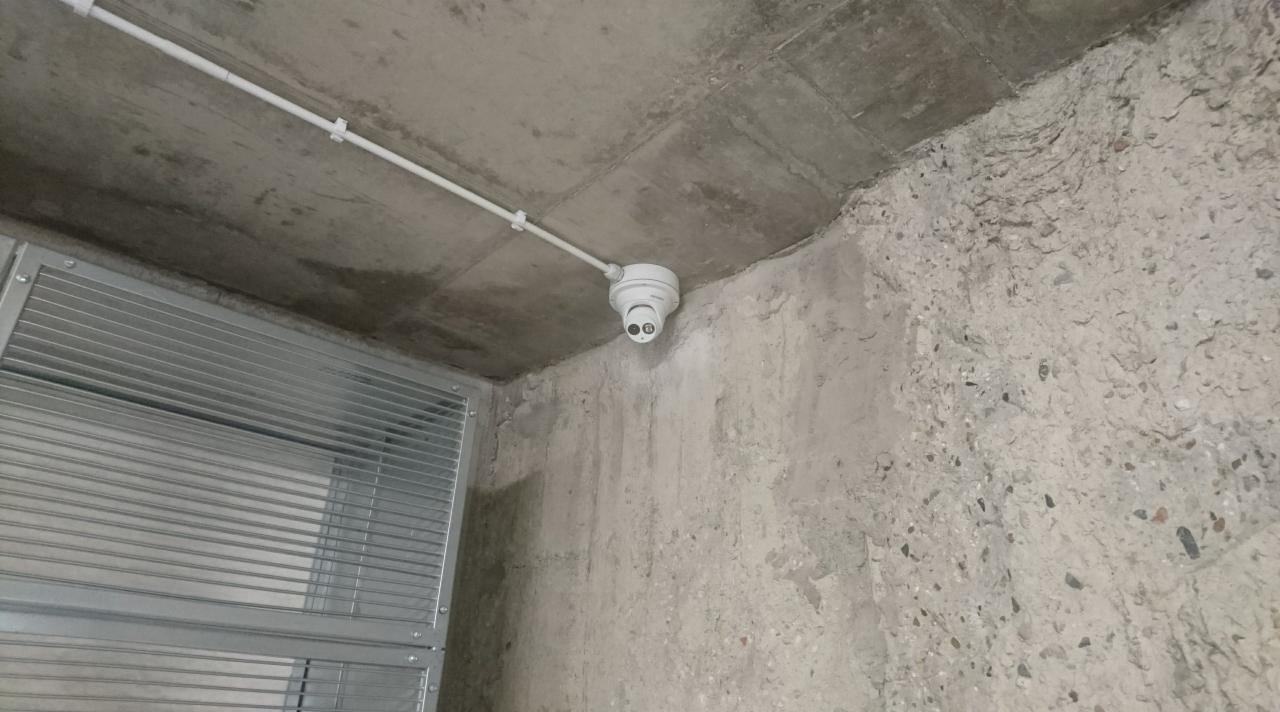 Kamera kopułkowa CCTV zamontowana na suficie w rogu pomieszczenia, widoczna instalacja kablowa w białej rurce, surowy beton na ścianach i suficie, metalowa żaluzja po lewej stronie.