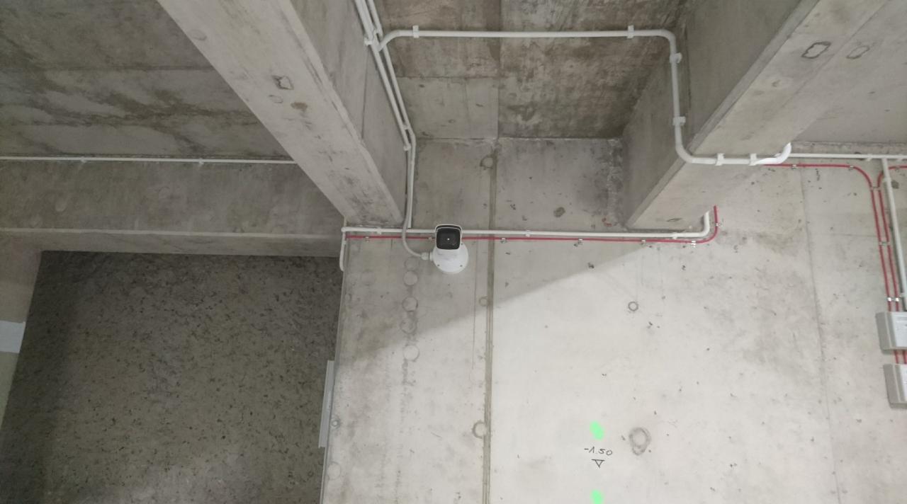 Kamera monitoringu zamontowana na betonowej ścianie z widocznymi przewodami elektrycznymi w białych i czerwonych rurkach. Widoczne zielone oznaczenia laserowe na ścianie.