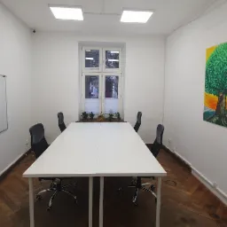 Jasne pomieszczenie biurowe z białym stołem konferencyjnym, krzesłami na kółkach, tablicą suchościeralną, zegarem, obrazem drzewa na ścianie i roślinami w oknie.