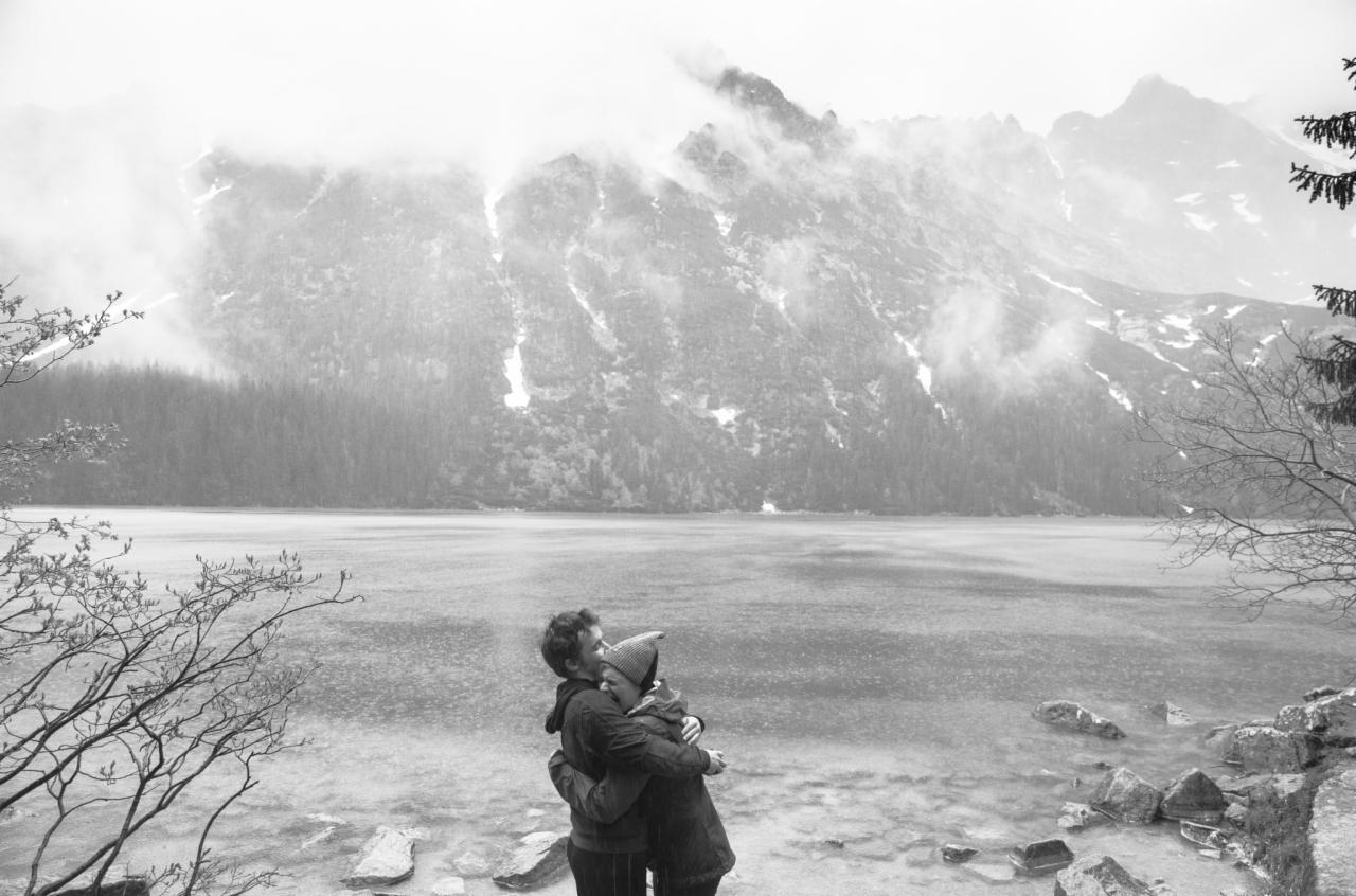 Czarno-białe zdjęcie pary obejmującej się nad brzegiem jeziora z widokiem na góry spowite mgłą, wykonane w pochmurny dzień.