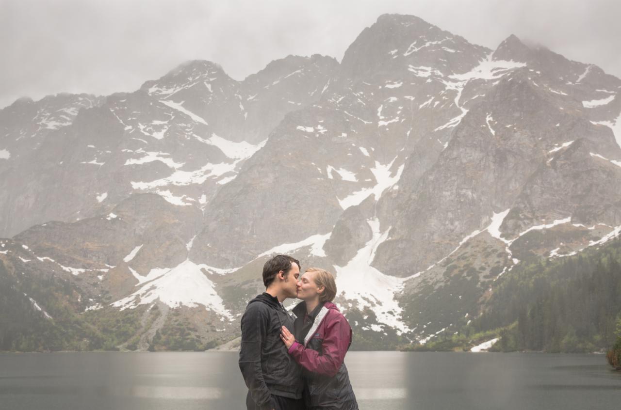 Para całująca się na tle górskiego jeziora w pochmurny dzień, kobieta w bordowej kurtce opiera dłoń na piersi mężczyzny