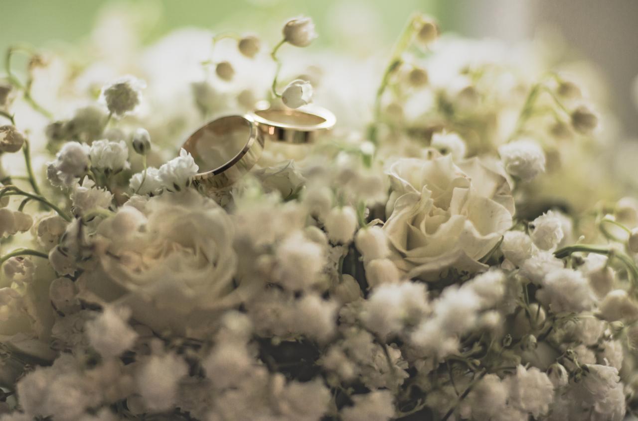 Dwa złote obrączki ślubne spoczywają na bukiecie z białych róż i konwalii, uchwycone w delikatnym, rozmytym świetle.