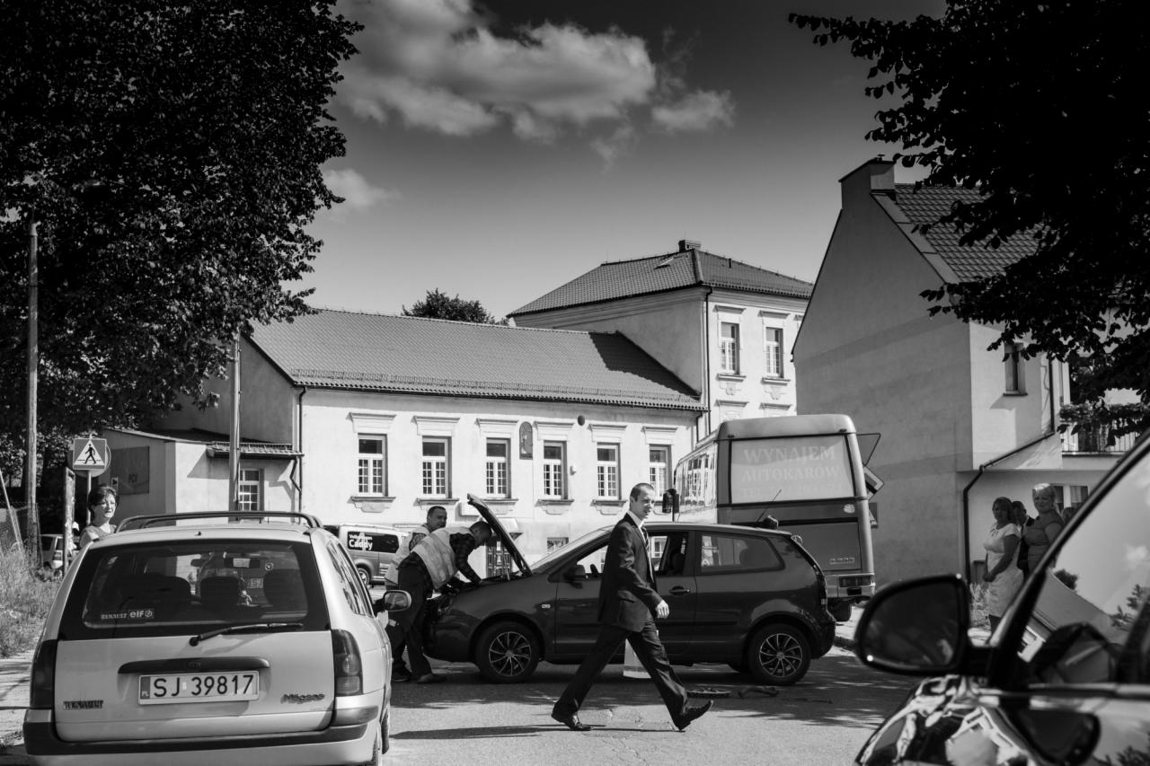 Czarno-biała fotografia reportażowa: mężczyzna w garniturze przechodzi obok otwartej maski samochodu, przy którym pracuje mechanik, w tle budynki i autobus.