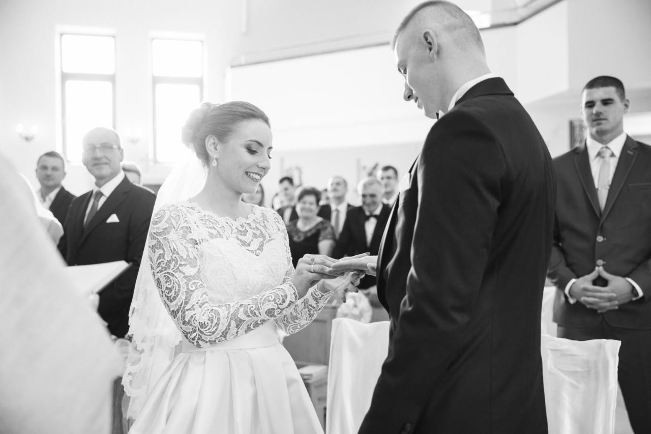 Czarno-białe zdjęcie z ceremonii ślubnej w kościele: uśmiechnięta panna młoda w koronkowej sukni zakłada obrączkę na palec pana młodego, w tle widoczni goście.