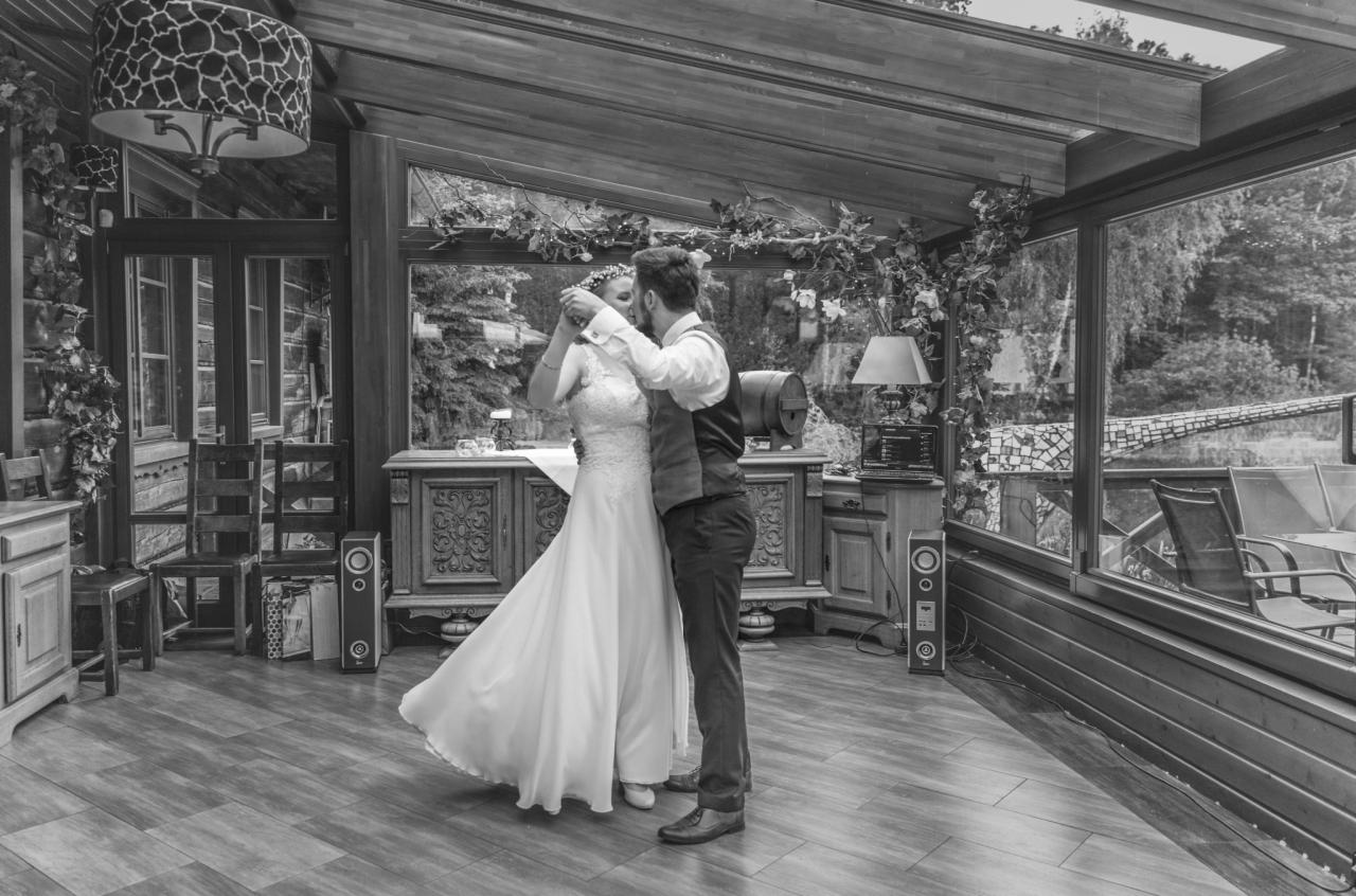 Czarno-białe zdjęcie pary młodej podczas pierwszego tańca w przeszklonej sali weselnej, dekoracje roślinne, drewniane meble i parkiet.