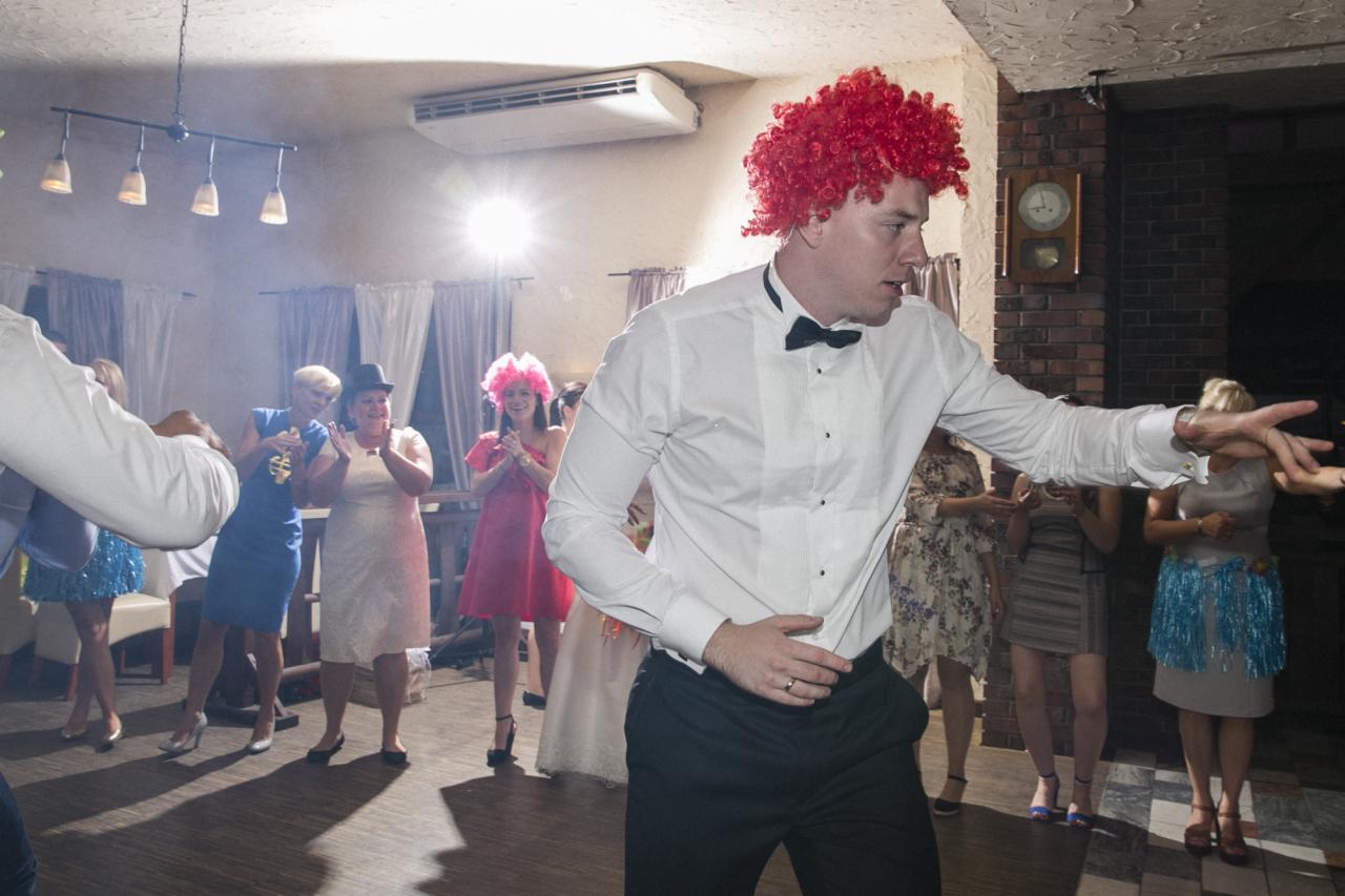 Mężczyzna w muszce i czerwonej peruce tańczy na weselu, w tle goście i dekoracje.