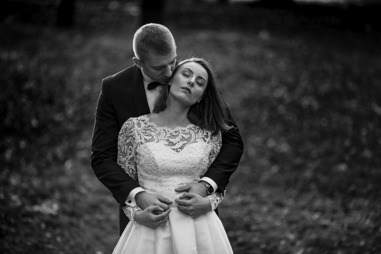 Czarno-białe zdjęcie pary młodej: pan młody w smokingu obejmuje pannę młodą w koronkowej sukni z zamkniętymi oczami, w tle rozmyty plener.