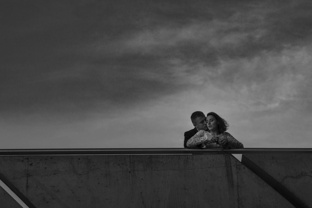 Czarno-białe zdjęcie pary w plenerze: mężczyzna w garniturze obejmuje kobietę w koronkowej sukience na tle pochmurnego nieba, opierając się o betonową balustradę.