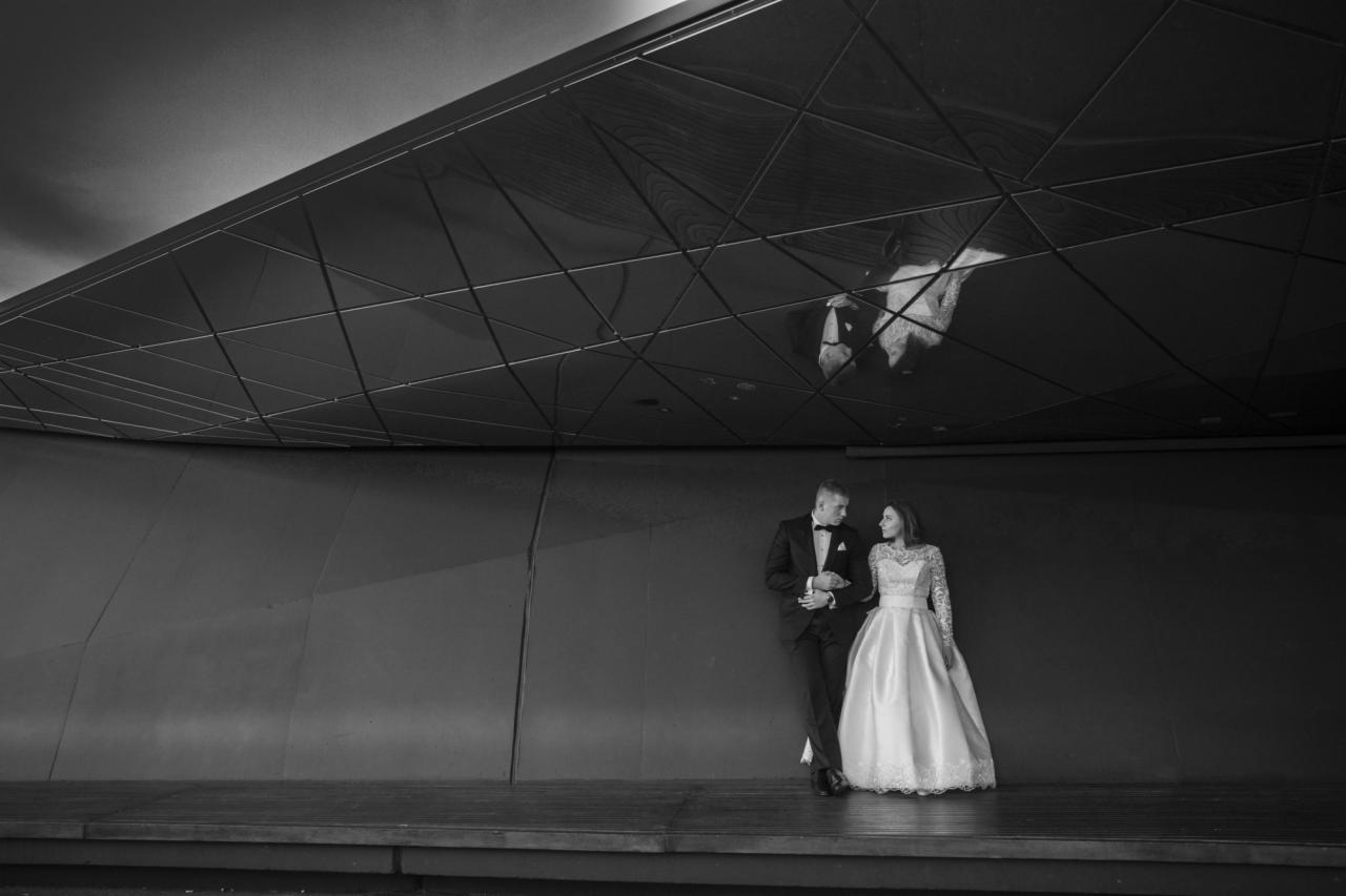 Czarno-białe zdjęcie pary młodej: pan młody w smokingu oparty o ścianę, panna młoda w koronkowej sukni spogląda na niego, w tle nowoczesna architektura z geometrycznym sufitem.