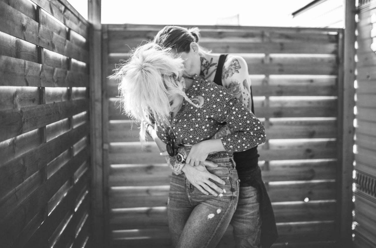 Czarno-białe zdjęcie pary obejmującej się na tle drewnianej ściany; kobieta z blond włosami w koszuli w kropki i jeansach, mężczyzna z tatuażami na ramionach i szyi.