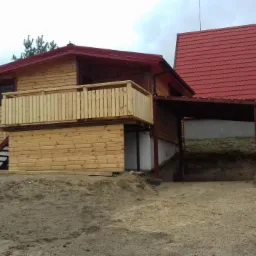 Drewniany dom z czerwonym dachem, widoczne elementy elewacji drewnianej, balkon z drewnianą balustradą oraz wiata garażowa. Teren wokół domu niezagospodarowany, piaszczysty.