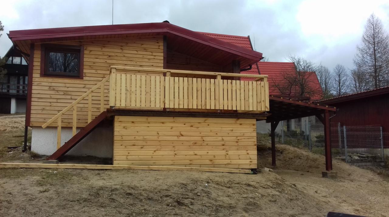 Nowo wybudowany, drewniany domek letniskowy z czerwonym dachem, balkonem i schodami, otoczony ziemią. Widoczna konstrukcja z drewna i fragmenty innych budynków w tle.